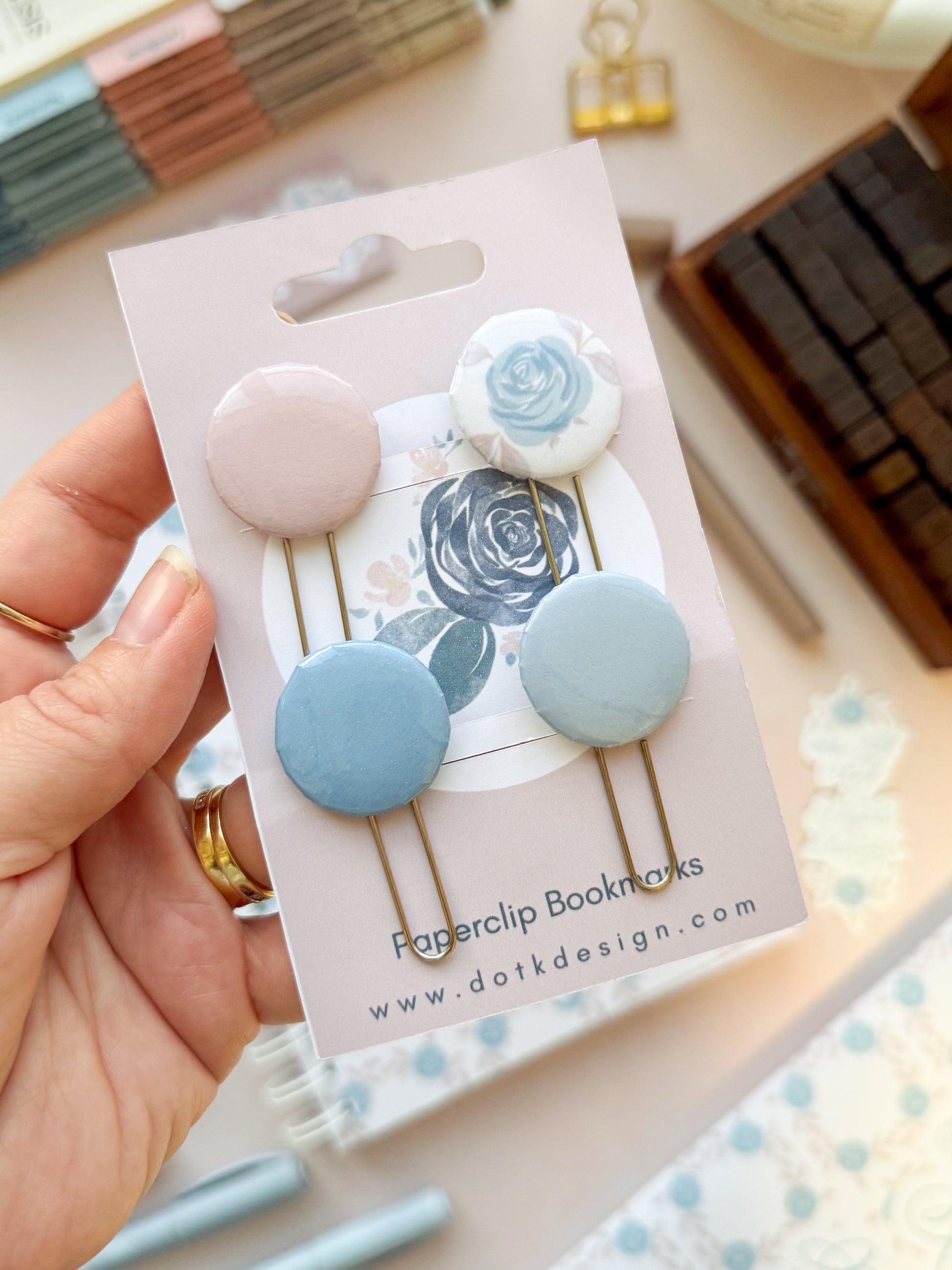 Hand holding a set of paperclip bookmarks with floral designs on a DotK Design branded card.