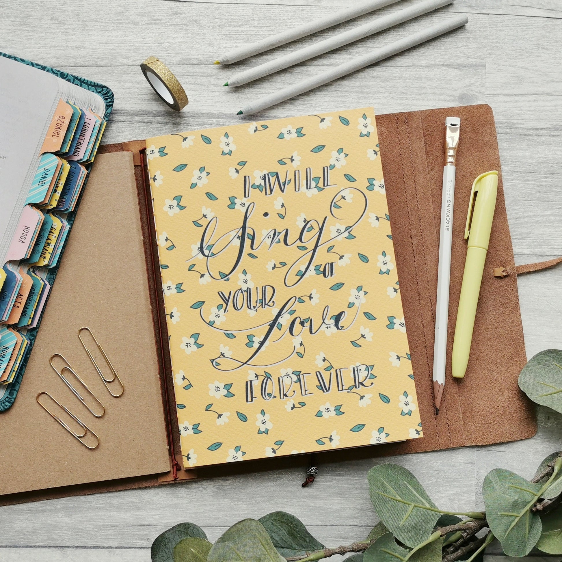 Christian prayer journal notebook in bright yellow with white daisy design and the word "I will sing of Your love forever" on the cover.